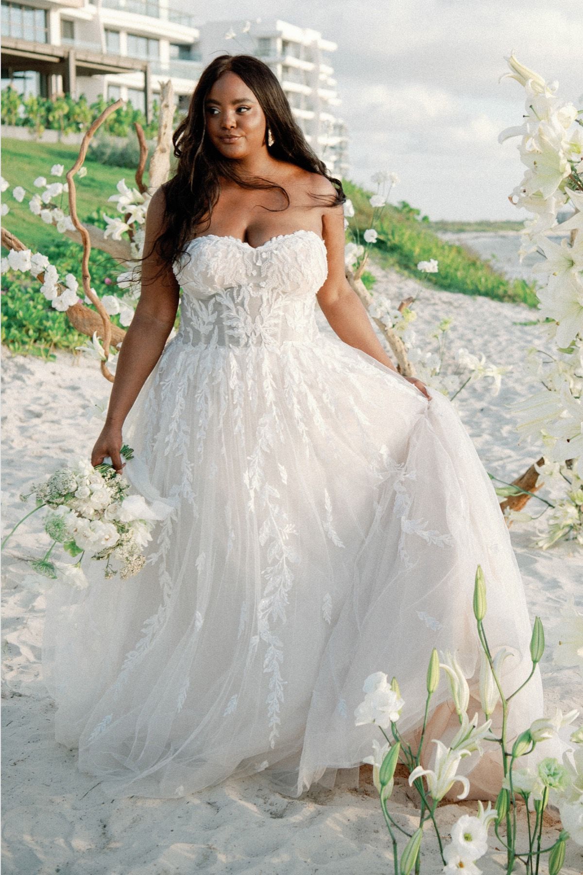 A woman in a wedding dress is standing on a beach holding a bouquet of flowers.