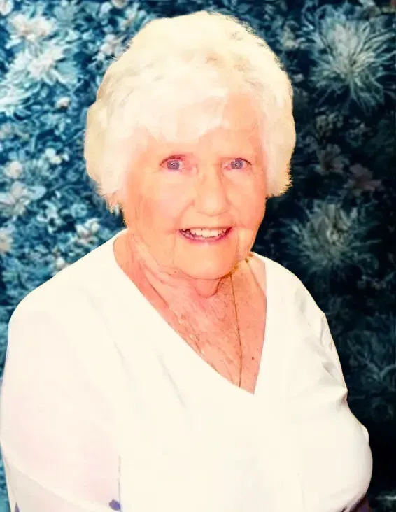 Smiling person with white hair wearing a white v-neck top against a blue floral background.