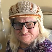 A person with blonde hair wears a crocheted tan and brown cap and black-framed glasses while facing forward.