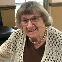 A smiling person with glasses, gray hair, and a pearl necklace, wearing a brown shirt and white crocheted cardigan.