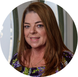 Headshot of a person with shoulder-length brown hair, wearing a patterned top, smiling at the camera.