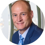 Headshot of a man with short hair wearing a blue suit and light blue tie against a blurred office background.