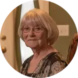 An older person with short gray hair and glasses, wearing a patterned top, looking toward the camera in a room.