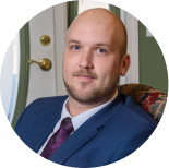 A professional headshot of a person wearing a blue suit and a dark patterned tie, set against an indoor backdrop.