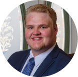 A person wearing a blue suit and patterned tie smiling in front of a decorative glass window.
