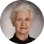 Head-and-shoulders portrait of an individual with short, light hair wearing a dark top against a gray background.