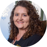 Headshot of a person with curly brown hair, wearing a dark blazer, smiling in front of a blue and white patterned backdrop.