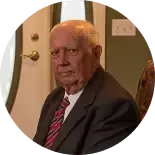A person wearing a suit and striped tie sits in an indoor setting with a door in the background.