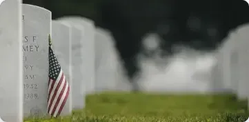 An American flag tucked against a white gravestone in a cemetery with rows of markers stretching into the distance.