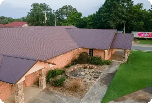 A high-angle view of a single-story brick building with a dark brown metal roof, stone pillars, and landscaped front.
