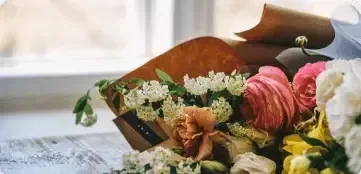 A bouquet of mixed pink, white, and yellow flowers wrapped in brown paper resting on a wooden surface by a window.