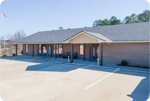 A single-story, light-colored brick commercial building with a gray shingled roof and a paved parking lot in the foreground.