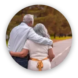 Two people with gray hair walk arm-in-arm along a road, viewed from behind. The person on the right wears a gold belt.