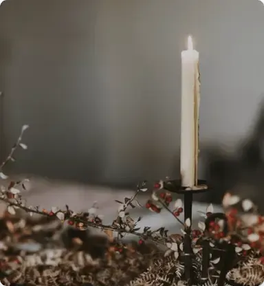 A lit white taper candle sits in a dark metal holder, surrounded by sprigs of dried greenery and red berries on a table.