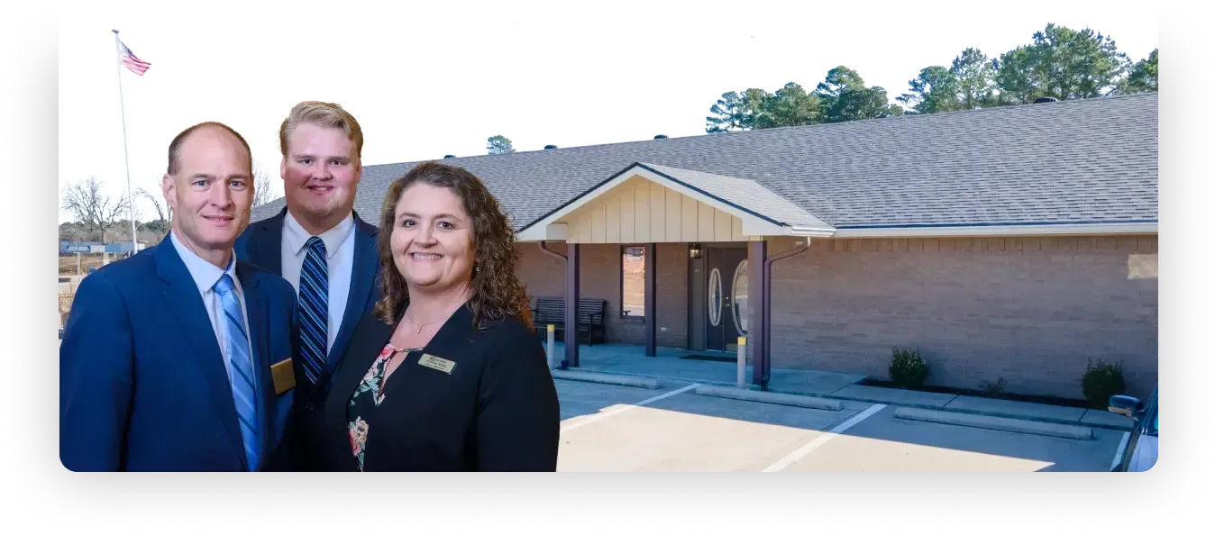 Three people in business attire stand in front of a brick building with a peaked entrance.