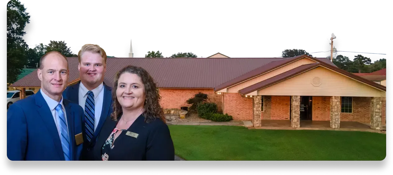 Three people in professional attire stand smiling in front of a brick building with a covered entrance and green lawn.