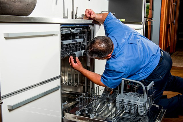 Man in blue shirt repairing a dishwasher, kneeling in front of it in a kitchen.