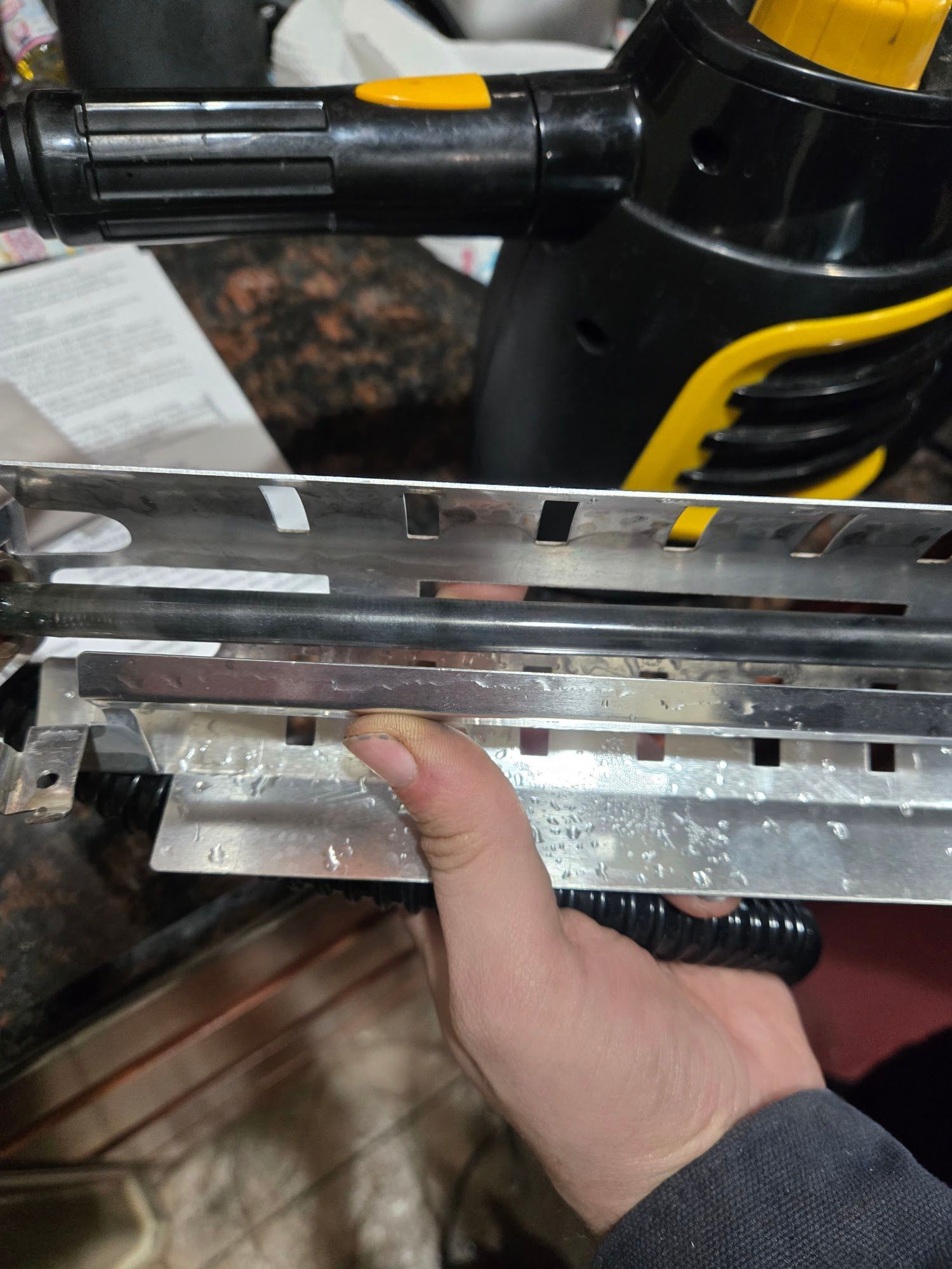 A hand holding a disassembled, silver and black steam cleaner attachment, next to the steam cleaner handle.