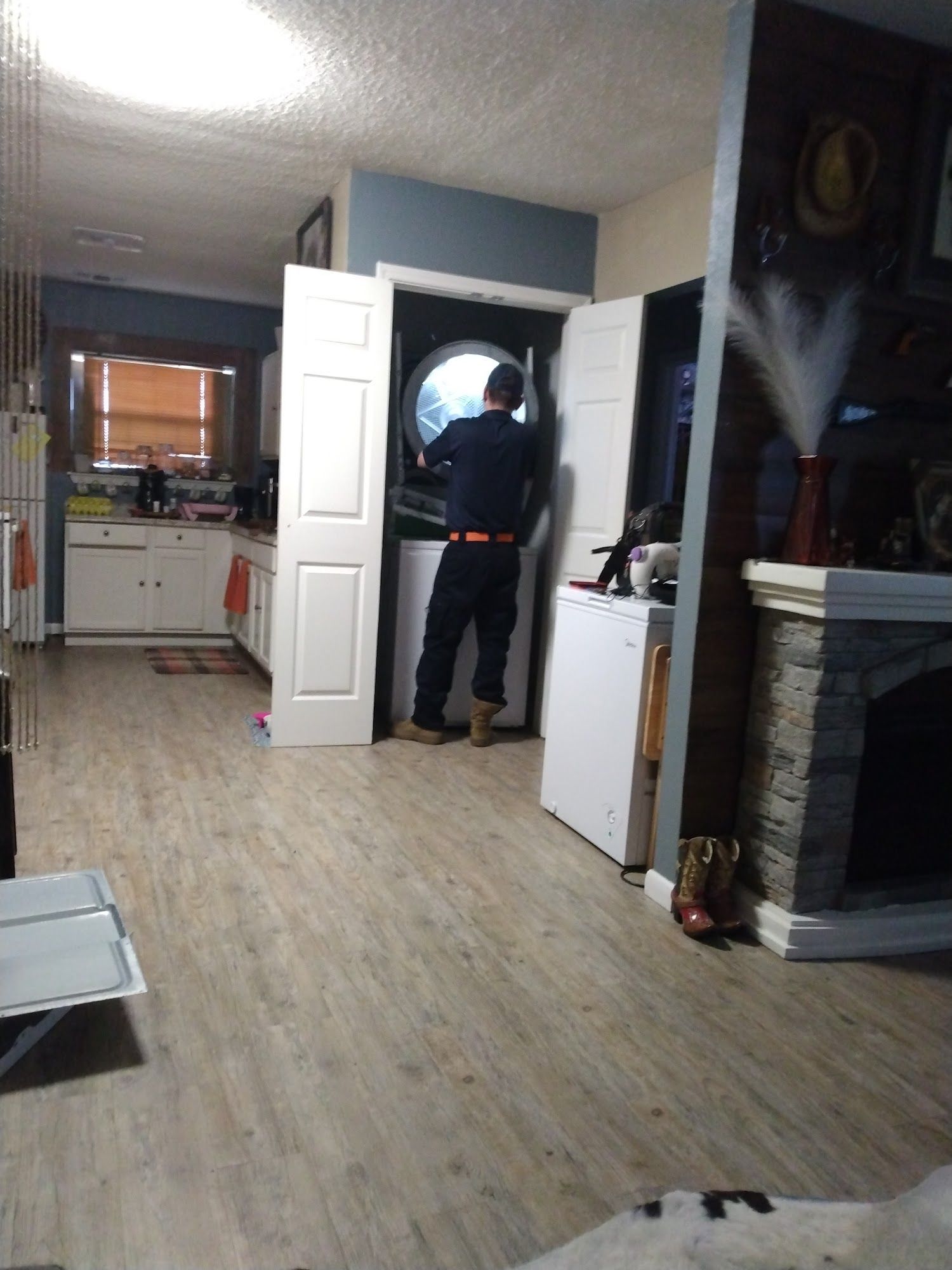 Person in dark clothing working on a washing machine in a laundry area. Light wooden floor, kitchen and fireplace visible.