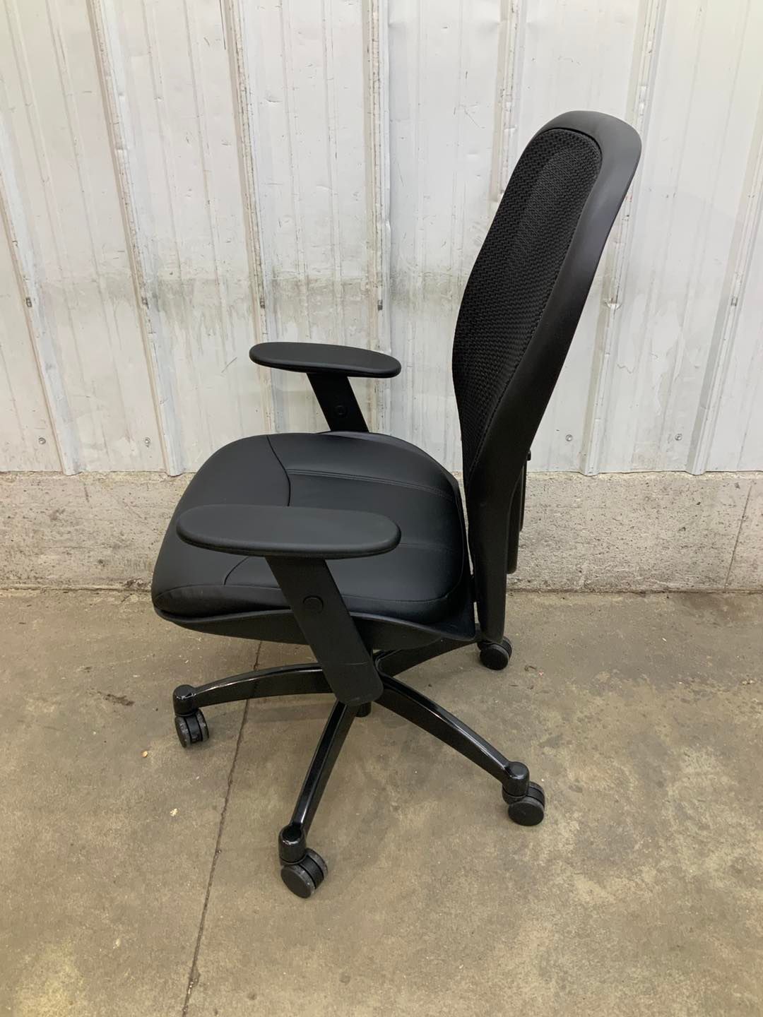 A black office chair is sitting on a concrete floor in front of a white wall.