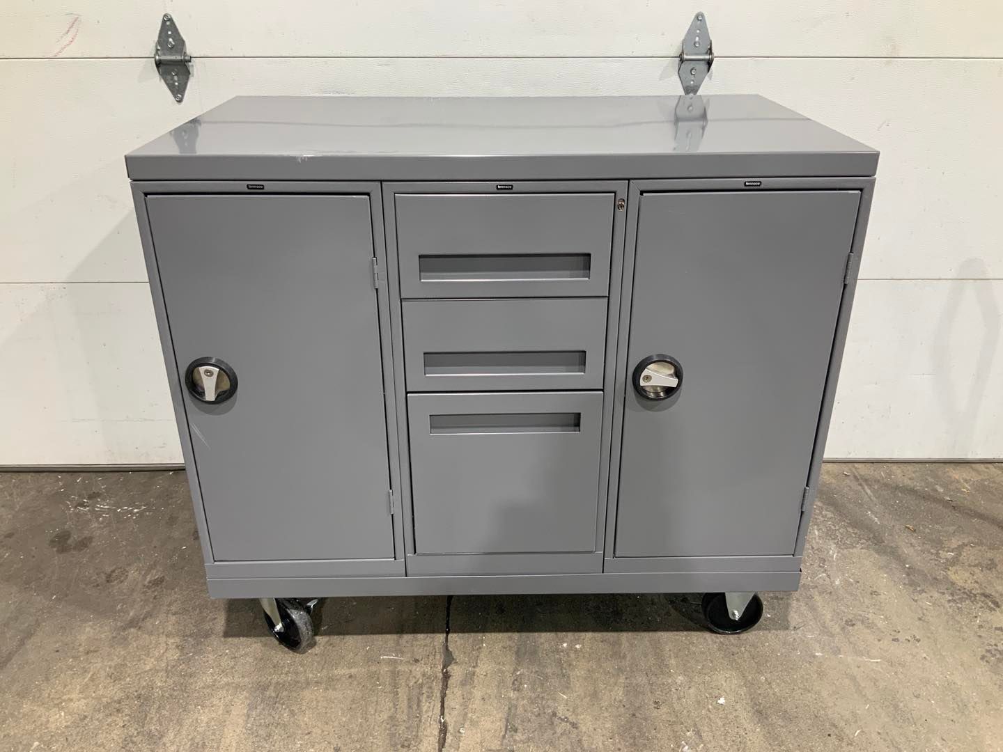 A gray metal cabinet with wheels is sitting on the ground in front of a garage door.