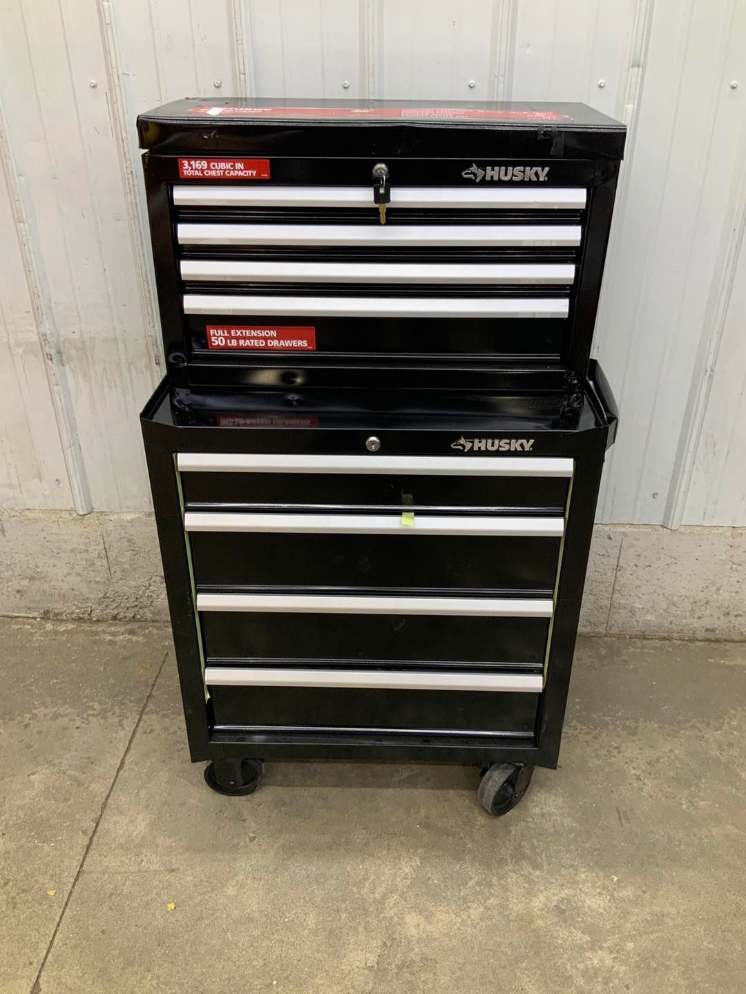 A black husky toolbox is sitting on the ground next to a white wall.