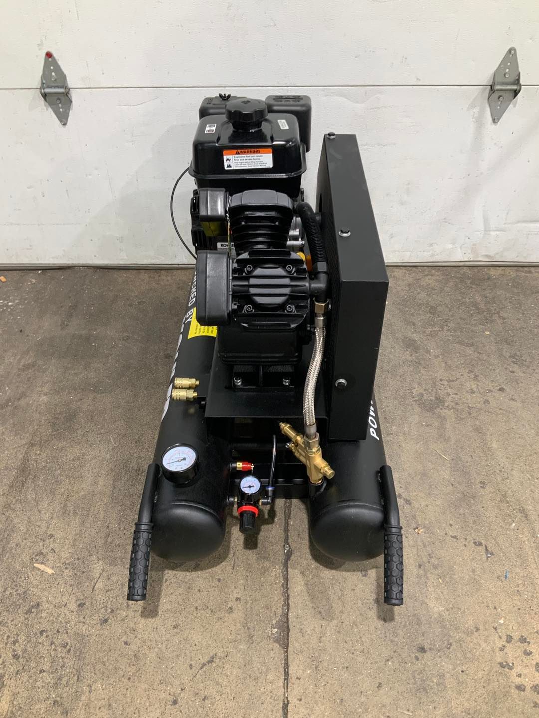 A black air compressor is sitting on the ground in front of a garage door.