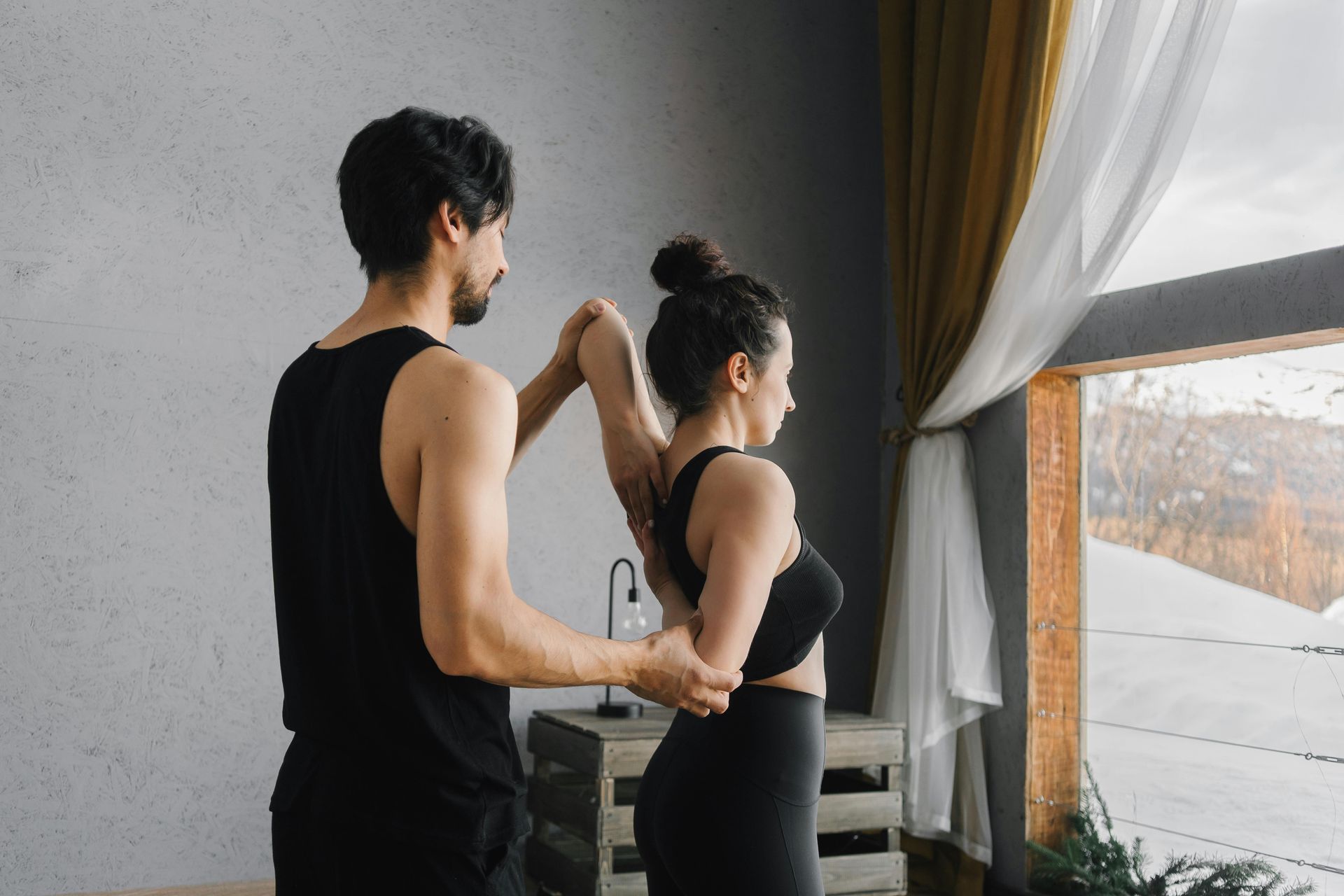 A man is helping a woman stretch her arms in front of a window.