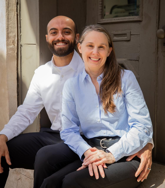 Couple sitting close together, smiling. Man in white shirt, woman in blue, both with black pants. Outdoor setting.