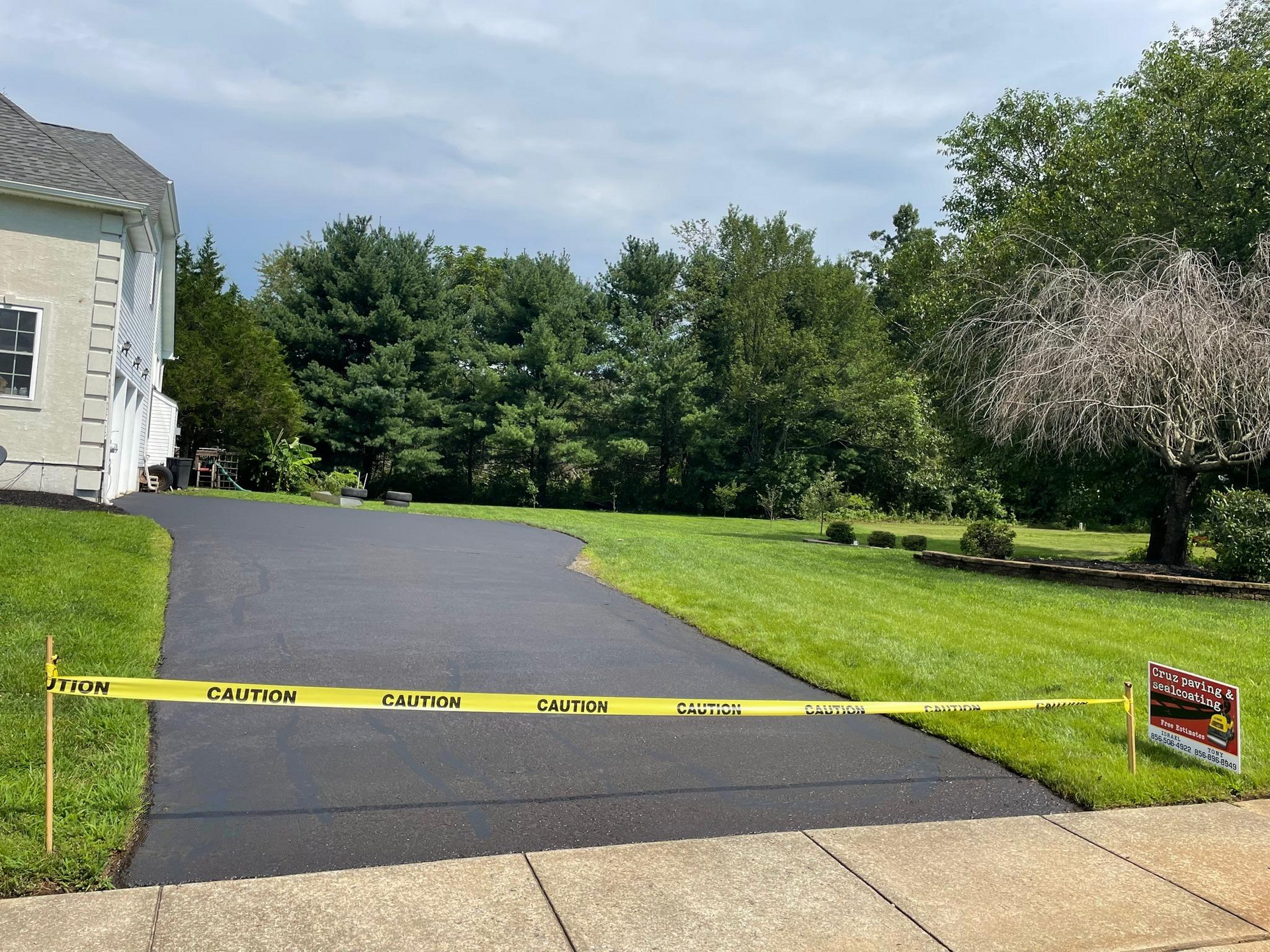 Yellow caution tape blocks a newly paved driveway beside a house and lawn, with a sign on the right