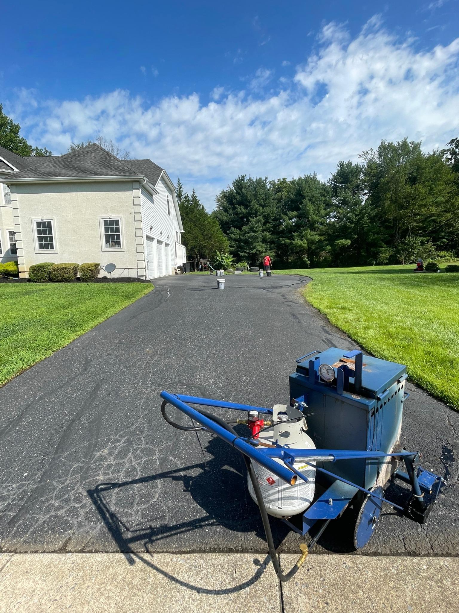 blue asphalt repair machine on a driveway beside a white house under a bright blue sky