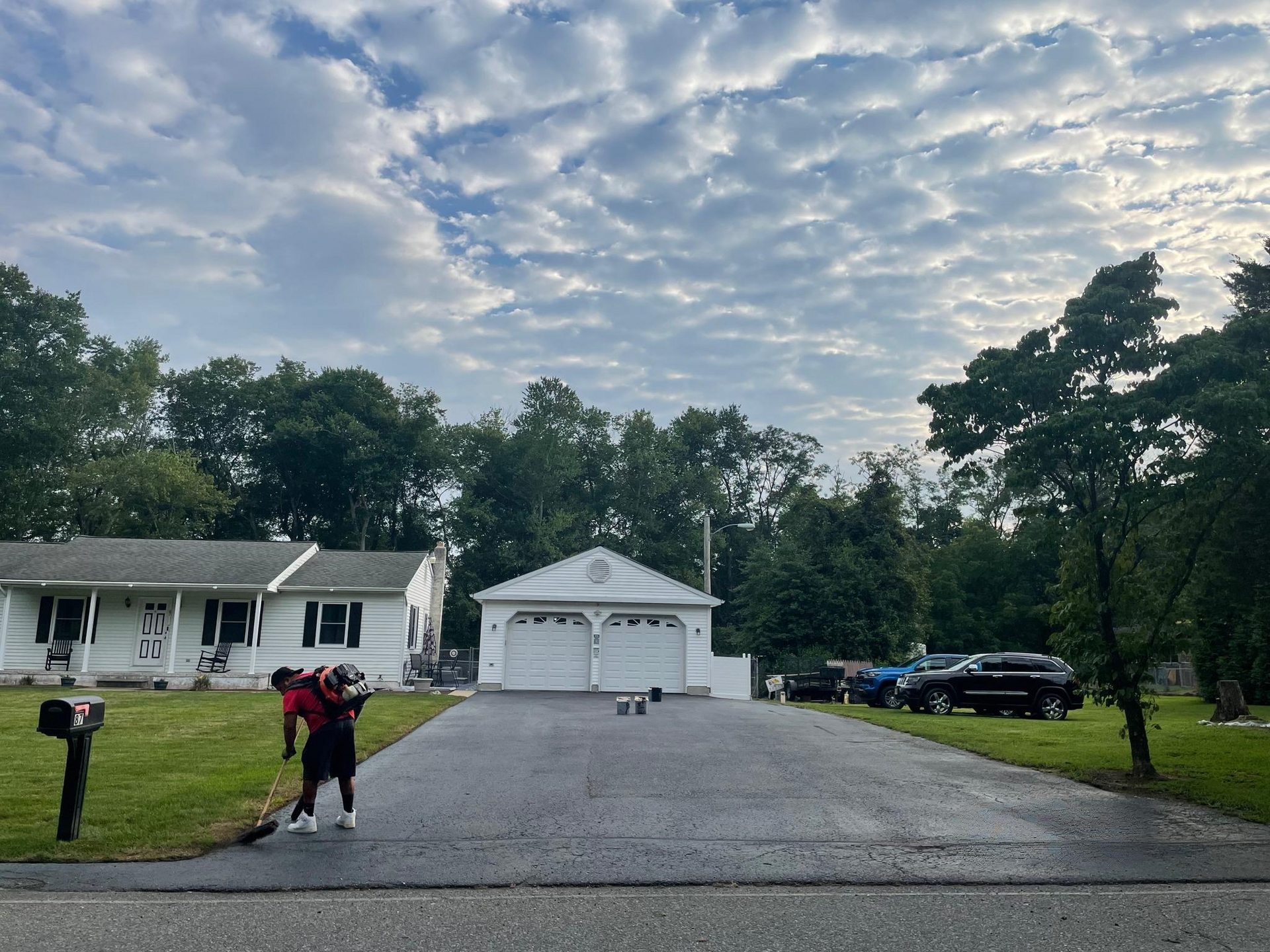 Suburban house with long driveway, trees, and two people walking near parked cars under a cloudy sky