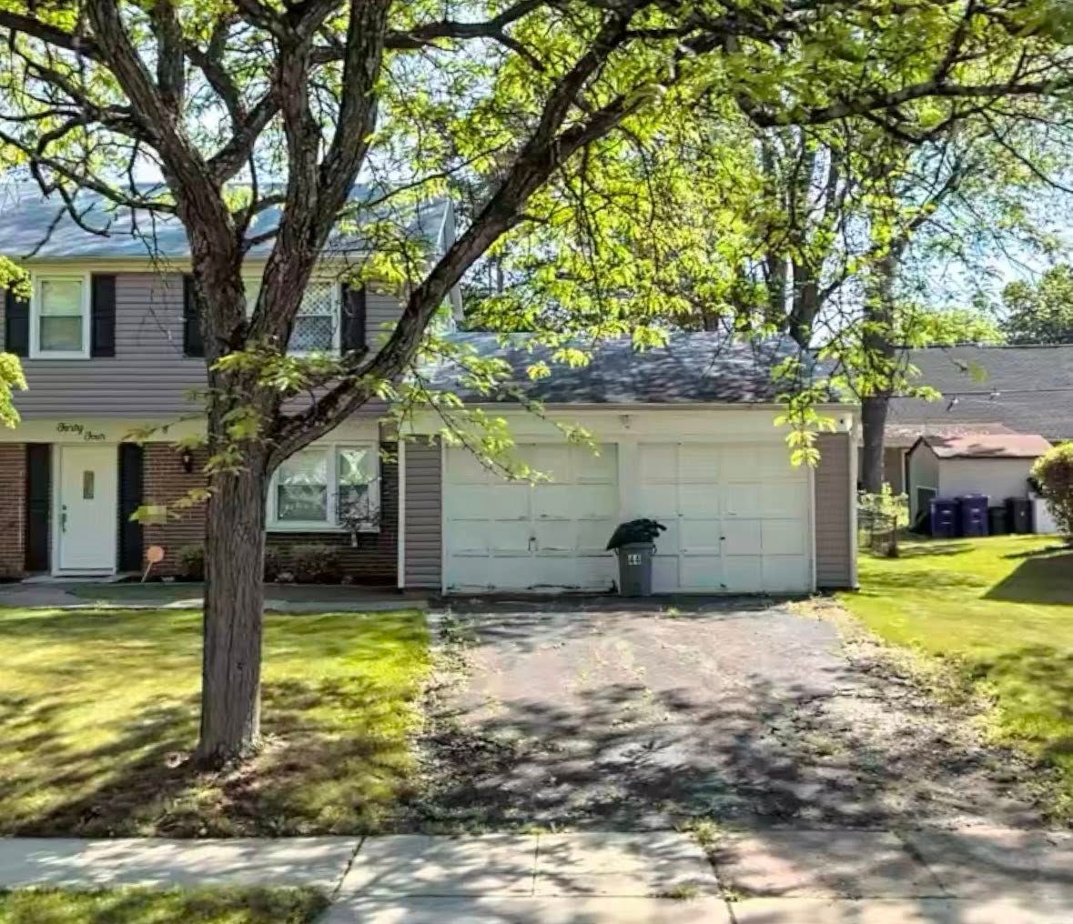 Suburban house with a two-car garage, tree-lined front yard, and driveway in sunlight
