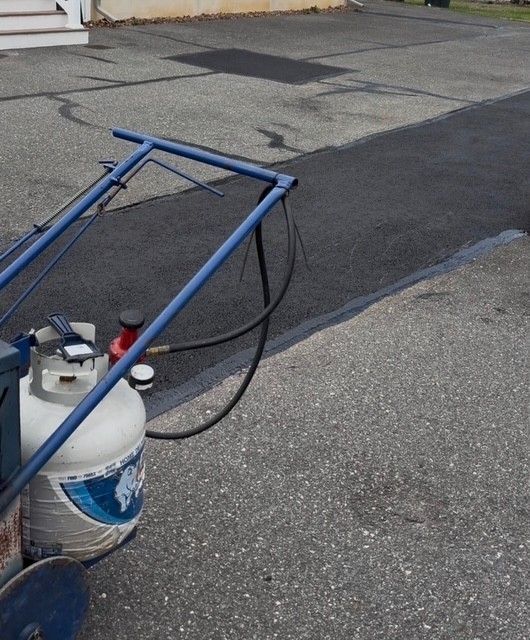 Blue spray rig with a propane tank on a paved driveway, spraying black sealant or paint onto asphalt