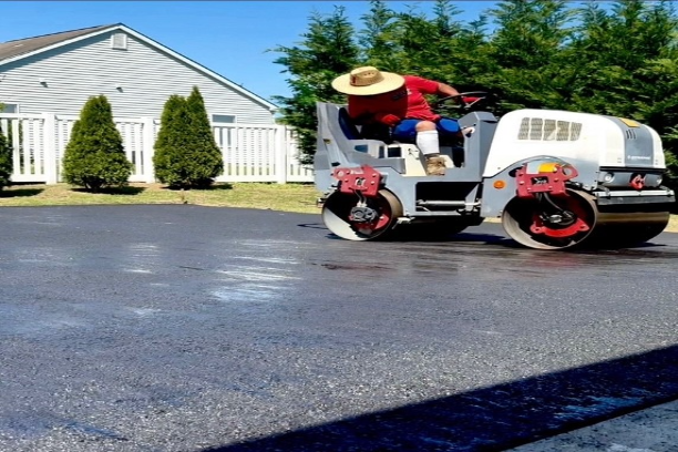 Cruz Paving & Sealcoating employee riding a white pavement rolling vehicle on a paved lot beside a white house