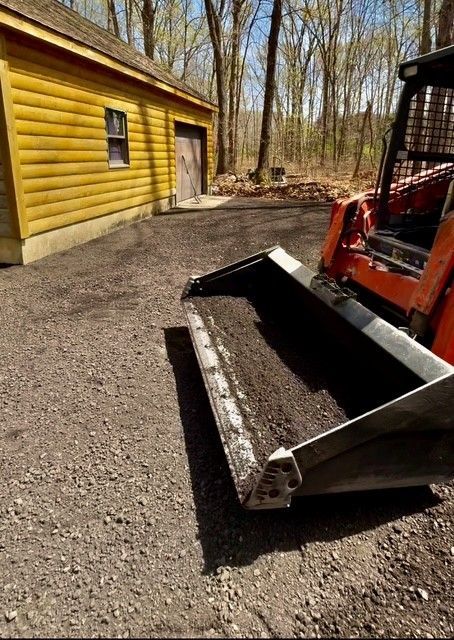 Crushed asphalt millings inside an orange skid-steer bucket on gravel beside yellow building in wooded area