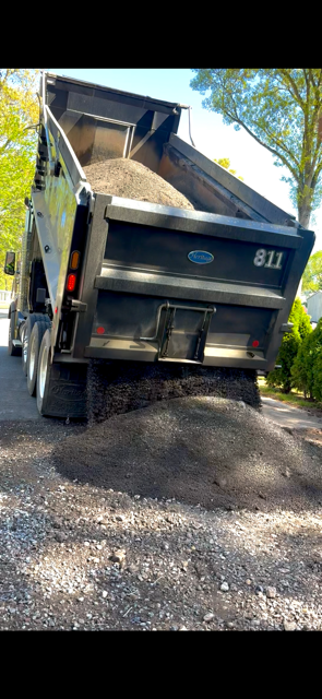 Dump truck unloading asphalt millings on a dirt driveway in a wooded area.