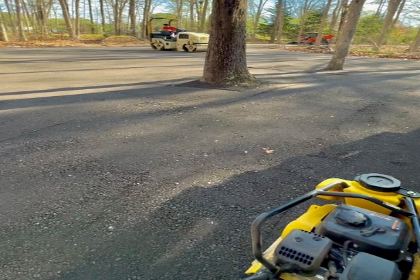Yellow paving compactor in the foreground of a masphalt millings parking lot with trees and another paving compactor in the background