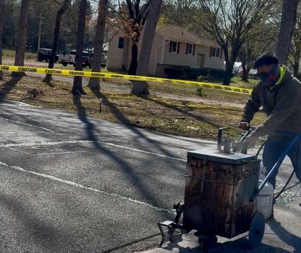 Cruz Paving & Sealcoating employee using a propane-powered asphalt repair tool beside a road near yellow caution tape and a house