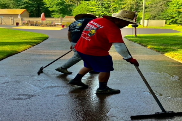 Two Cruz Paving & Sealcoating employees spreading sealcoating application on a sidewalk in a park, one in red and one in gray
