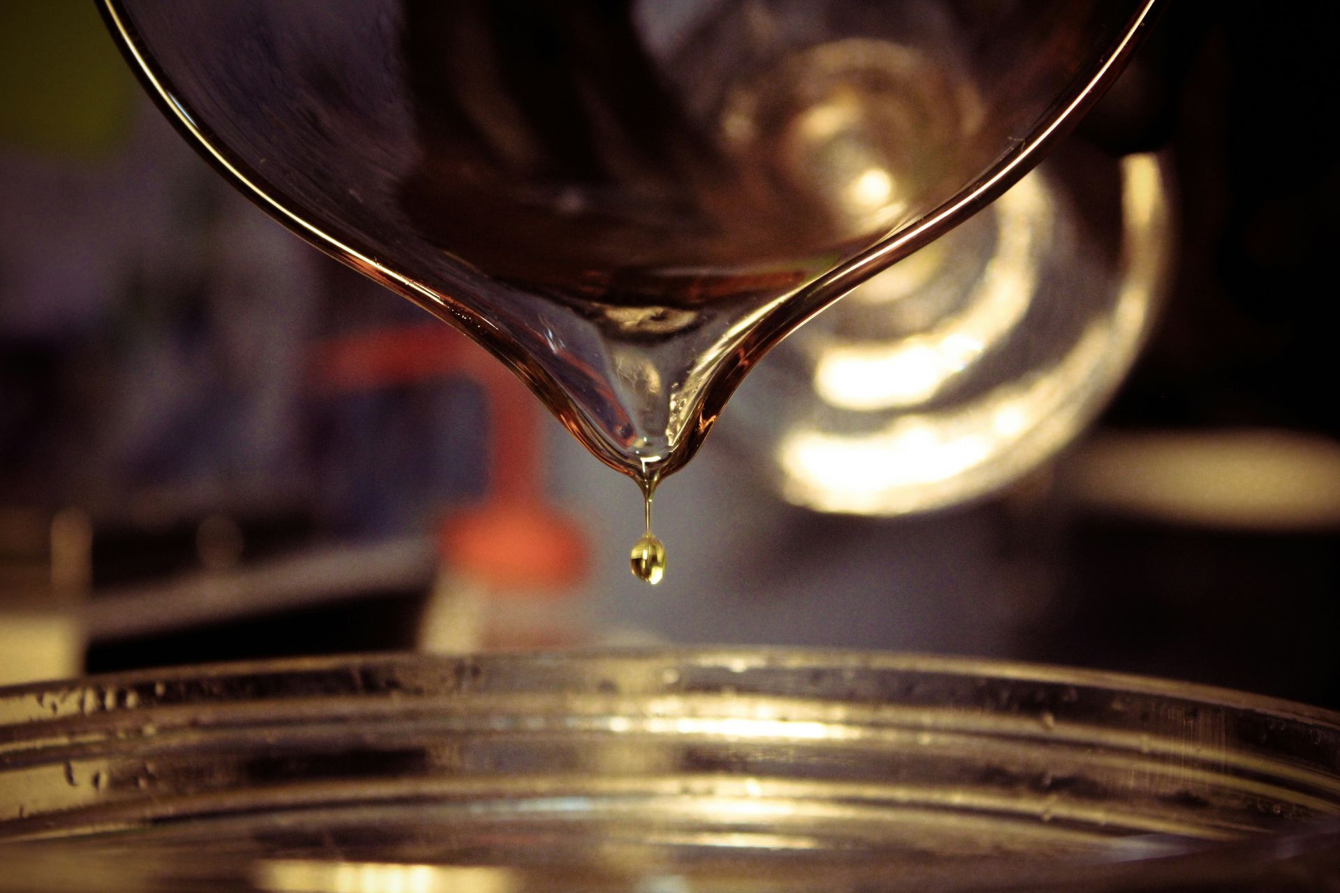Close-up of a silver pitcher pouring a golden liquid into a metal bowl, with a single drop hanging.