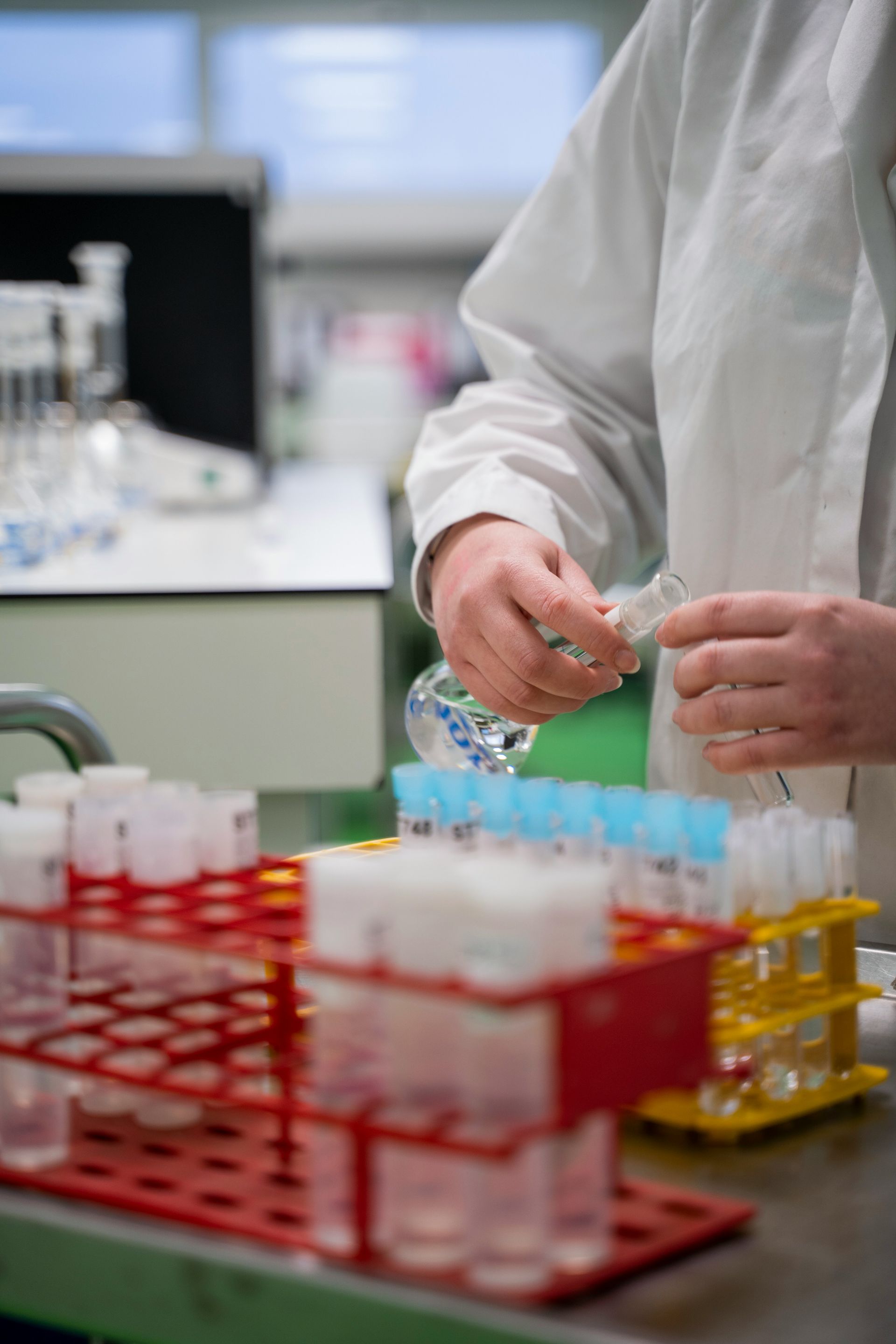 Person in lab coat handling vial with beakers and test tubes in the background.