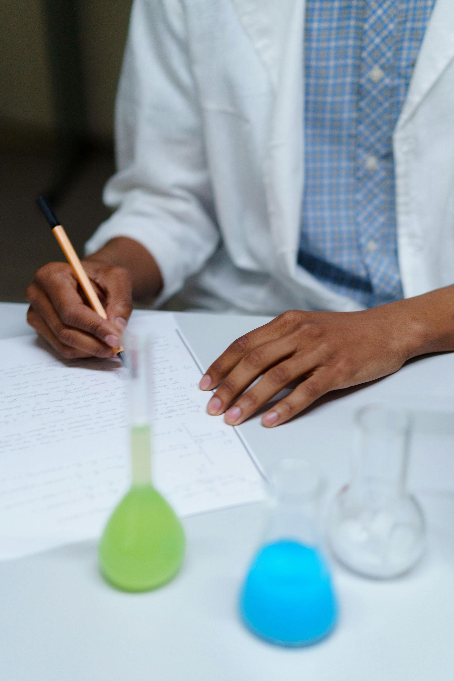 Scientist in lab coat writing notes with beakers of green, blue, and clear liquid on the table.