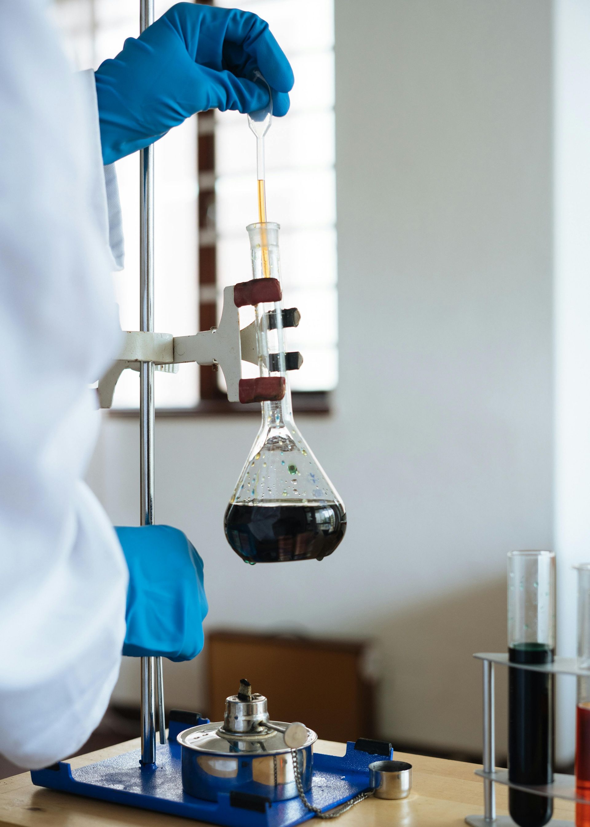 Scientist in lab coat and gloves performing titration with dark solution in a flask, lab setting.