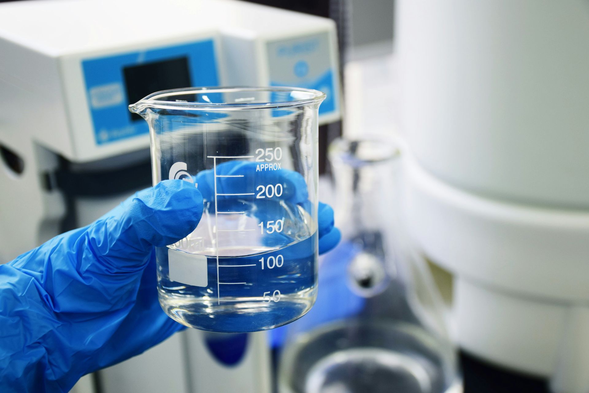 Hand in blue glove holds a beaker of clear liquid in a lab.