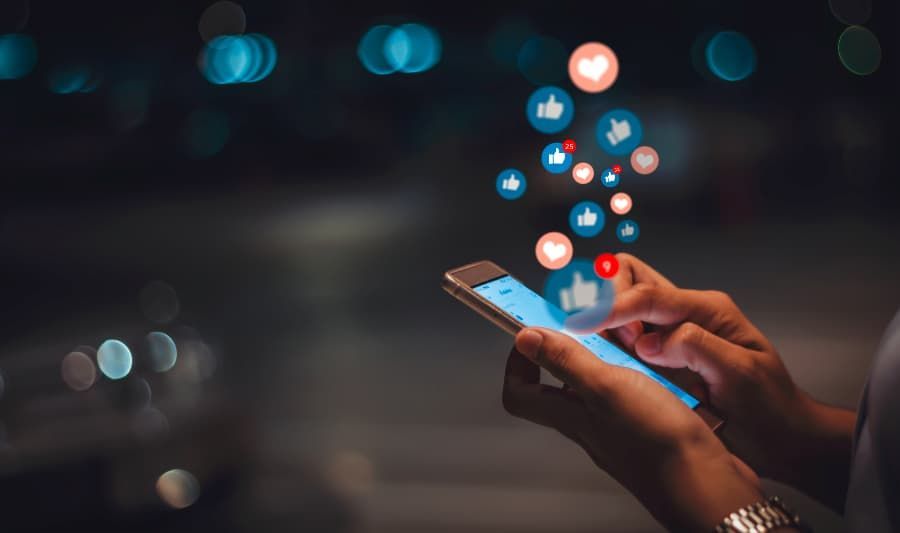 Hands holding a smartphone with social media emojis coming out the top. 