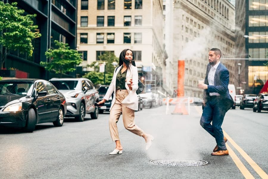 Professional man and woman running past a steaming manhole cover, jaywalking in NYC.