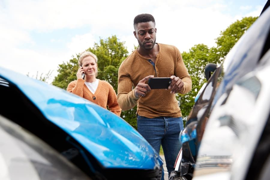 Man taking photos of wrecked vehicles, with woman on phone behind him. 