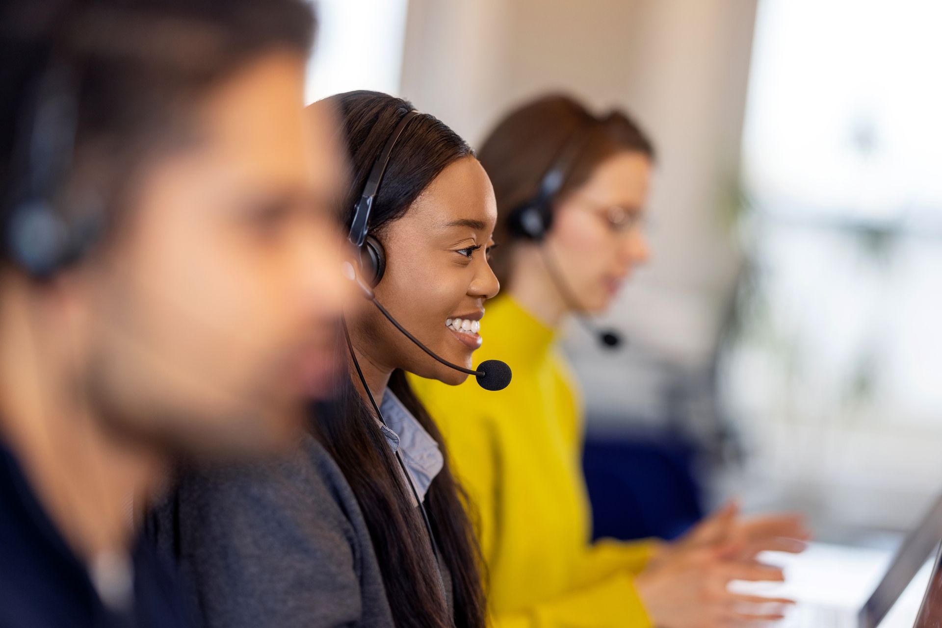 Row of call center agents wearing headsets and working at computers in a bright office.
