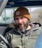 A man with a beard and a beanie is sitting in the driver 's seat of a car.